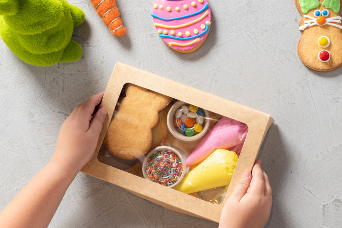 Easter-themed cookies and decorations on a gray surface with a child's hands holding a box of cookie ingredients.